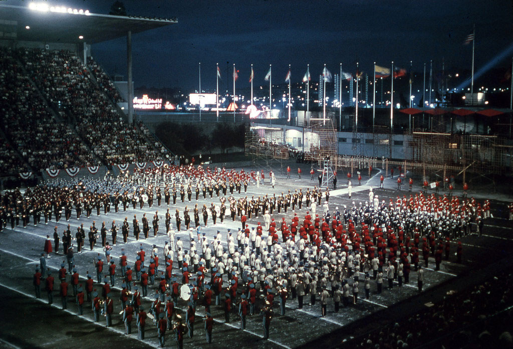 A color photograph of red and white dressed marching bands in the stadium at the Seattle Center at night.  The stands are full of people and spotlights hit the band and many nations flags fly in the background. 
